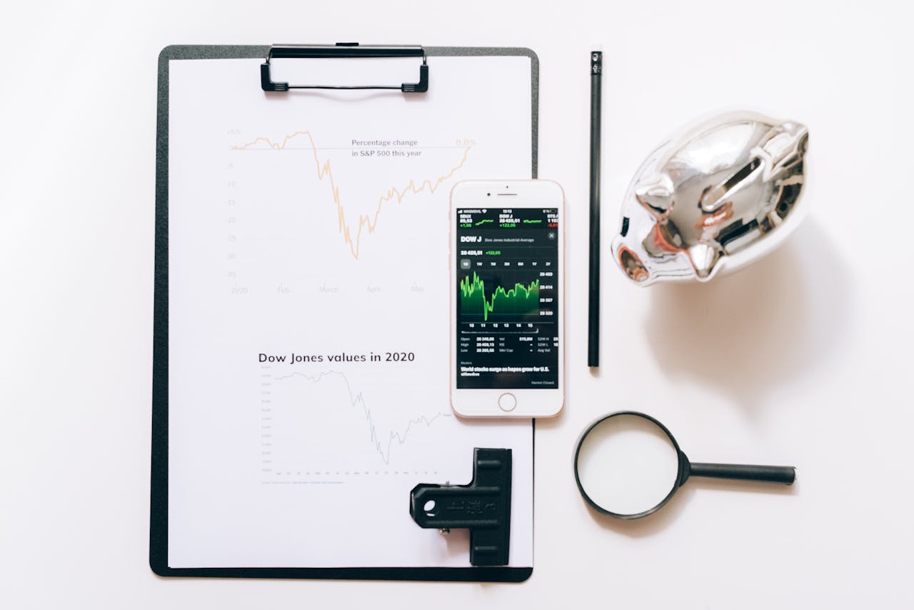 Top view of financial analysis tools including a cellphone, clipboard, magnifying glass, and piggy bank on a white desk.